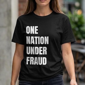 May include: A black short-sleeved t-shirt featuring the white text ONE NATION UNDER FRAUD printed in a bold, sans-serif font across the chest. The garment has a classic crew neckline and a straight hem, providing a simple and clean design.