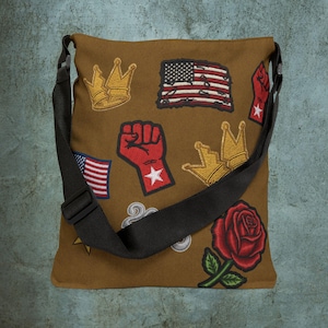 Op de afbeelding: Bruine canvas schoudertas met een verstelbare zwarte riem. De tas is versierd met geborduurde patches, waaronder Amerikaanse vlaggen, gouden kronen, een rode vuist, een rode roos en een ster.