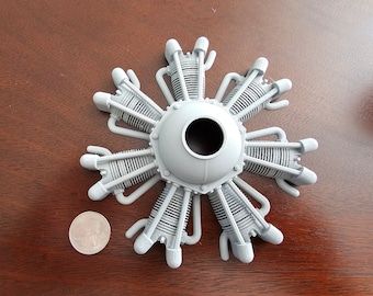Puede incluir: Un modelo de motor radial de 9 cilindros de color gris. El motor tiene un cubo central con una abertura circular y ocho cilindros que irradian hacia afuera. El modelo está hecho de plástico y tiene un diámetro de aproximadamente 7,5 cm.
