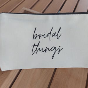 Könnte beinhalten: Eine weiße Canvas-Reißverschlusstasche mit schwarzem Besatz und dem aufgedruckten Text "bridal things" in Schwarz.