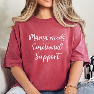 K&ouml;nnte beinhalten: Ein weinrotes T-Shirt mit dem wei&szlig;en Text "Mama needs Emotional Support". Das Shirt hat kurze &Auml;rmel und eine lockere Passform. Das Model tr&auml;gt ein goldenes Armband mit blauen Akzenten.