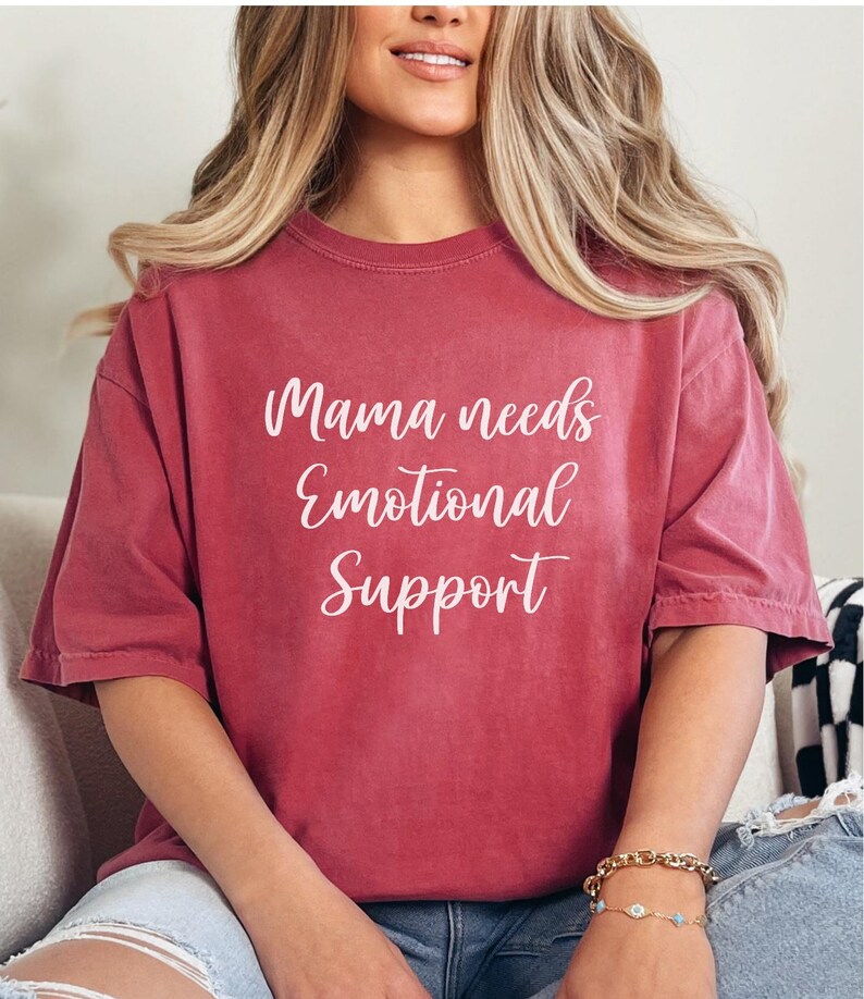 K&ouml;nnte beinhalten: Ein weinrotes T-Shirt mit dem wei&szlig;en Text "Mama needs Emotional Support". Das Shirt hat kurze &Auml;rmel und eine lockere Passform. Das Model tr&auml;gt ein goldenes Armband mit blauen Akzenten.