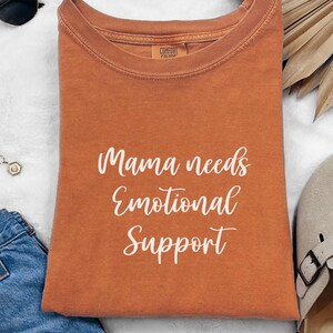 K&ouml;nnte beinhalten: Ein orangefarbenes Comfort Colors T-Shirt mit dem wei&szlig;en Schriftzug "Mama needs Emotional Support". Das Shirt ist gefaltet und hat einen Rundhalsausschnitt. Das Bild enth&auml;lt Sonnenbrillen, eine Halskette und eine Jeans.