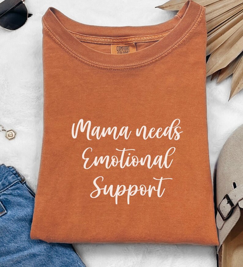 K&ouml;nnte beinhalten: Ein orangefarbenes Comfort Colors T-Shirt mit dem wei&szlig;en Schriftzug "Mama needs Emotional Support". Das Shirt ist gefaltet und hat einen Rundhalsausschnitt. Das Bild enth&auml;lt Sonnenbrillen, eine Halskette und eine Jeans.