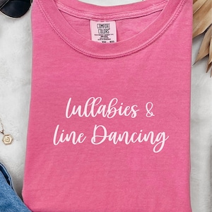 May include: Pink t-shirt with white text that reads "Lullabies & Line Dancing".