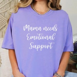 K&ouml;nnte beinhalten: Helllila T-Shirt mit wei&szlig;em Schriftzug "Mama needs Emotional Support". Das Oberteil hat kurze &Auml;rmel und ist einfarbig.