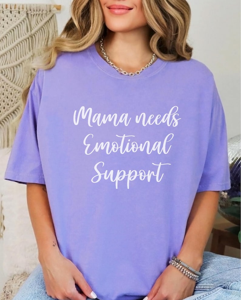 K&ouml;nnte beinhalten: Helllila T-Shirt mit wei&szlig;em Schriftzug "Mama needs Emotional Support". Das Oberteil hat kurze &Auml;rmel und ist einfarbig.