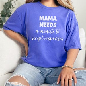 May include: A blue t-shirt with the text "MAMA NEEDS a minute to script responses" in white. The shirt is a solid color and has short sleeves. The person is wearing blue jeans and a gold bracelet.