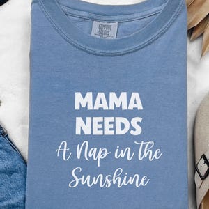 May include: A light blue t-shirt with the text "Mama Needs A Nap in the Sunshine" printed in white.
