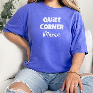 May include: A blue t-shirt with the words "QUIET CORNER Mama" in white block and script font. The shirt has short sleeves and a crew neck. The model is wearing light blue jeans and a gold bracelet.