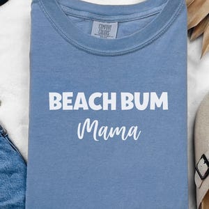 May include: A light blue t-shirt with the text "Beach Bum Mama" in white lettering.