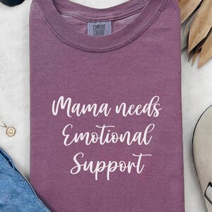 K&ouml;nnte beinhalten: Ein mauvefarbenes Comfort Colors T-Shirt mit dem wei&szlig;en Schriftzug "Mama needs Emotional Support". Das Shirt ist gefaltet, mit einem Etikett am Halsausschnitt. Sonnenbrille und Halskette sind im Hintergrund zu sehen.
