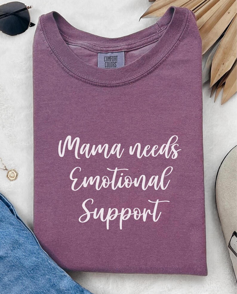 K&ouml;nnte beinhalten: Ein mauvefarbenes Comfort Colors T-Shirt mit dem wei&szlig;en Schriftzug "Mama needs Emotional Support". Das Shirt ist gefaltet, mit einem Etikett am Halsausschnitt. Sonnenbrille und Halskette sind im Hintergrund zu sehen.