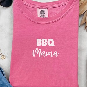 K&ouml;nnte beinhalten: Pinkes T-Shirt mit dem Text "BBQ Mama" in wei&szlig;en Buchstaben.