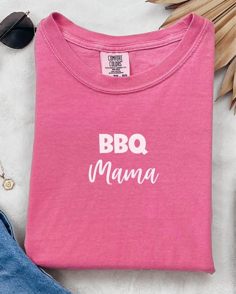 K&ouml;nnte beinhalten: Pinkes T-Shirt mit dem Text "BBQ Mama" in wei&szlig;en Buchstaben.