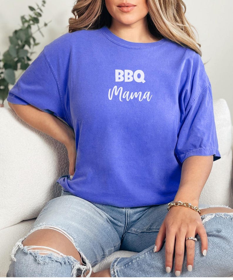 K&ouml;nnte beinhalten: Eine Frau tr&auml;gt ein lila T-Shirt mit dem Text "BBQ Mama" in Wei&szlig;. Sie tr&auml;gt eine blaue Jeans und ein goldenes Armband.