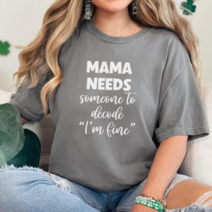 May include: Gray t-shirt with the text "MAMA NEEDS someone to decode 'I'm fine'" in white. The shirt is a relaxed fit, with short sleeves. The shirt is paired with blue jeans.