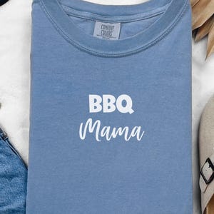 K&ouml;nnte beinhalten: Ein hellblaues T-Shirt mit dem Text "BBQ Mama" in wei&szlig;en Buchstaben. Das Shirt ist von Comfort Colors.