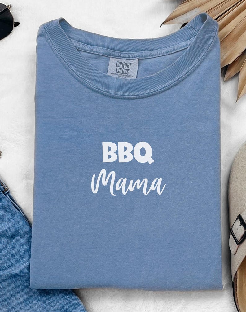 K&ouml;nnte beinhalten: Ein hellblaues T-Shirt mit dem Text "BBQ Mama" in wei&szlig;en Buchstaben. Das Shirt ist von Comfort Colors.