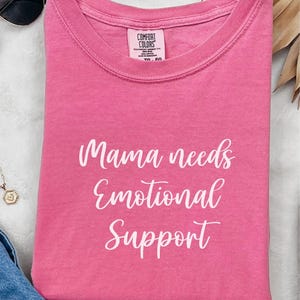 K&ouml;nnte beinhalten: Ein gefaltetes pinkfarbenes T-Shirt mit dem wei&szlig;en Schriftzug "Mama needs Emotional Support". Das T-Shirt hat einen Rundhalsausschnitt und ist aus weichem Material. Sonnenbrille und Halskette sind im Hintergrund zu sehen.
