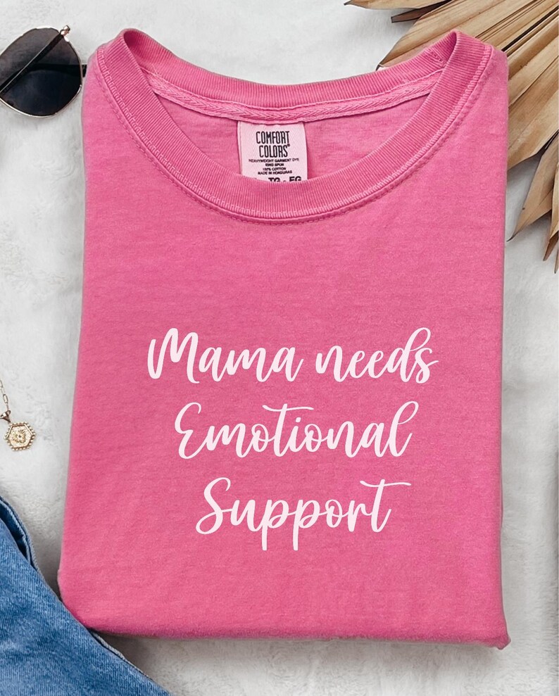 K&ouml;nnte beinhalten: Ein gefaltetes pinkfarbenes T-Shirt mit dem wei&szlig;en Schriftzug "Mama needs Emotional Support". Das T-Shirt hat einen Rundhalsausschnitt und ist aus weichem Material. Sonnenbrille und Halskette sind im Hintergrund zu sehen.