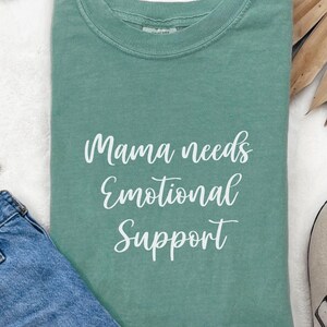 K&ouml;nnte beinhalten: Ein salbeigr&uuml;nes T-Shirt mit dem wei&szlig;en Schriftzug "Mama needs Emotional Support". Das Shirt ist gefaltet, eine blaue Jeans und eine Sonnenbrille sind zu sehen. Das T-Shirt hat einen Rundhalsausschnitt.