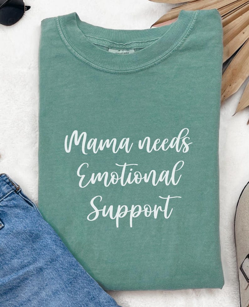 K&ouml;nnte beinhalten: Ein salbeigr&uuml;nes T-Shirt mit dem wei&szlig;en Schriftzug "Mama needs Emotional Support". Das Shirt ist gefaltet, eine blaue Jeans und eine Sonnenbrille sind zu sehen. Das T-Shirt hat einen Rundhalsausschnitt.
