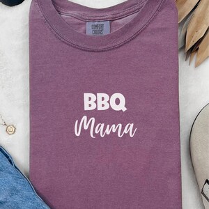 K&ouml;nnte beinhalten: Ein lila T-Shirt mit dem Text "BBQ Mama" in wei&szlig;en Buchstaben.