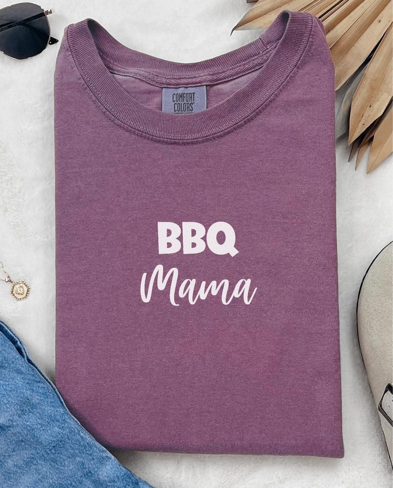 K&ouml;nnte beinhalten: Ein lila T-Shirt mit dem Text "BBQ Mama" in wei&szlig;en Buchstaben.