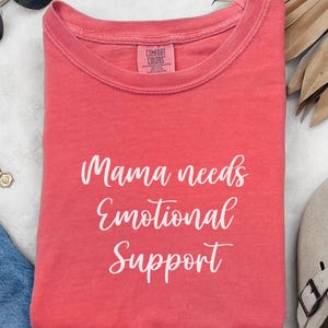K&ouml;nnte beinhalten: Gefaltetes korallenfarbenes T-Shirt mit dem wei&szlig;en Schriftzug "Mama needs Emotional Support". Das T-Shirt hat ein Comfort Colors-Etikett. Ein l&auml;ssiges Kleidungsst&uuml;ck.