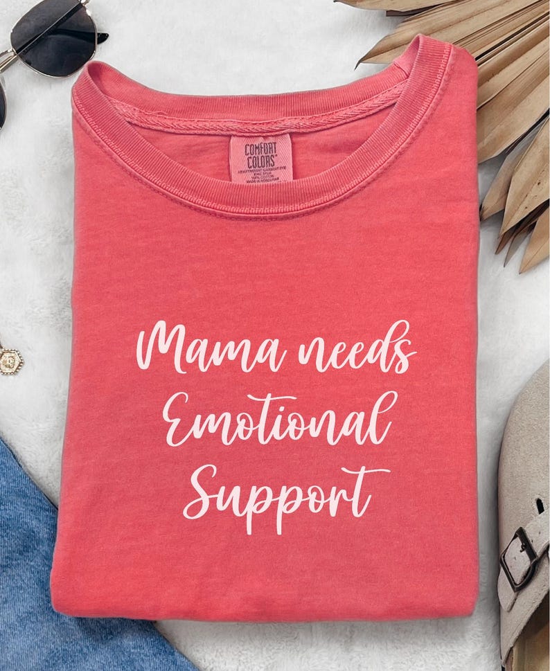 K&ouml;nnte beinhalten: Gefaltetes korallenfarbenes T-Shirt mit dem wei&szlig;en Schriftzug "Mama needs Emotional Support". Das T-Shirt hat ein Comfort Colors-Etikett. Ein l&auml;ssiges Kleidungsst&uuml;ck.