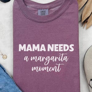 May include: A purple Comfort Colors t-shirt with the text "MAMA NEEDS a margarita moment" in white. The shirt is folded and displayed with sunglasses, a necklace, and a shoe.