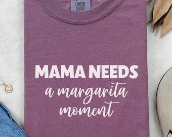 May include: A purple Comfort Colors t-shirt with the text "MAMA NEEDS a margarita moment" in white. The shirt is folded and displayed with sunglasses, a necklace, and a shoe.