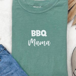 K&ouml;nnte beinhalten: Ein blaugr&uuml;nes T-Shirt mit dem Aufdruck "BBQ Mama" in Wei&szlig; auf der Vorderseite.