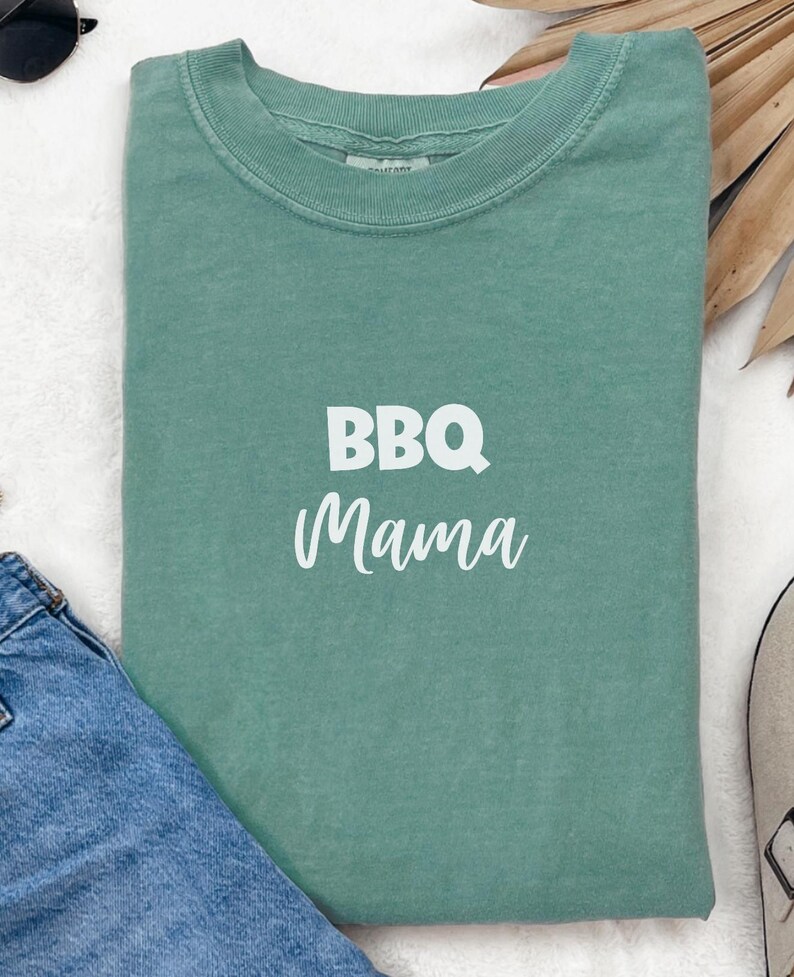 K&ouml;nnte beinhalten: Ein blaugr&uuml;nes T-Shirt mit dem Aufdruck "BBQ Mama" in Wei&szlig; auf der Vorderseite.