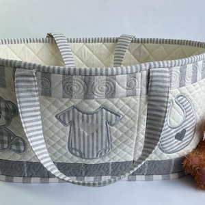 May include: A quilted fabric basket in cream and gray with striped handles and trim. The exterior features embroidered appliqués of a teddy bear, a baby bodysuit, and a bib. A small, fuzzy brown teddy bear sits next to the basket.