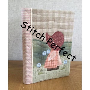 May include: A pink and beige quilted book cover featuring a girl in a red hat and a pink dress holding flowers. The text "Stitch Perfect" is on the cover.