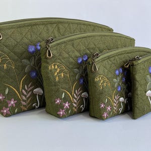 May include: A set of four olive green quilted zippered pouches in graduated sizes, each featuring embroidered botanical designs including small white mushrooms, purple flowers, blue clusters, and golden wheat stalks with bronze-toned zipper pulls.