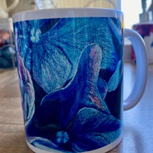 Puede incluir: Taza de cerámica blanca con un vibrante diseño floral azul. La obra de arte presenta flores azules detalladas sobre un fondo oscuro, con un asa y borde blancos. Adecuada para bebidas frías o calientes.