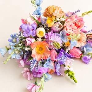 以下が含まれることがあります： ピーチ、ラベンダー、ライトブルー、イエローの色合いの造花をミックスした鮮やかなフラワーアレンジメント。ポピー、チューリップ、ラベンダーの小枝など、さまざまな花が含まれており、密集した華やかな花束を形成しています。