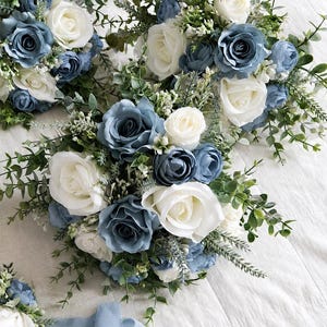 Puede incluir: Una colección de ramos de flores con rosas en azul polvoriento y blanco, acentuados con pequeñas flores blancas de relleno y hojas de eucalipto verdes. Los arreglos están atados con una cinta de tela azul claro, creando un conjunto floral.