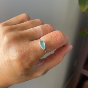 Puede incluir: Un anillo de plata con una piedra azul claro en forma de lágrima. El anillo tiene una banda delgada y un engaste de bisel. La piedra es de un color azul pastel suave y parece lisa y pulida. Una joya delicada.