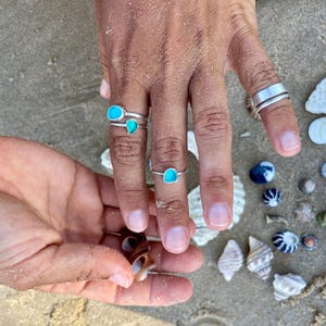 May include: Three silver rings with turquoise stones on a hand. The rings are on the index, middle, and ring fingers. The hand is resting on a surface with seashells.