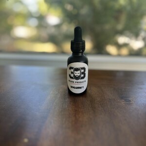 May include: A black glass bottle with a dropper top and a white label. The label has a black and white illustration of a man with a beard and the text "Beard Products Volume 1".