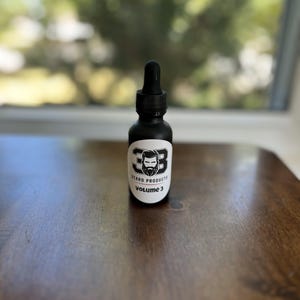 May include: A black glass bottle with a black dropper top and a white label. The label has a black and white illustration of a bearded man and the text "Beard Products Volume 3".