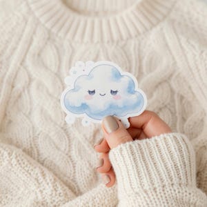 Könnte beinhalten: Ein gestanzter Aufkleber mit einer Aquarellillustration einer lächelnden Wolke in Blau und Weiß. Die Wolke hat rosige Wangen und geschlossene Augen. Kleine Sterne und Punkte umgeben die Wolke. Der Aufkleber wird vor einem cremefarbenen Strickpullover gehalten.