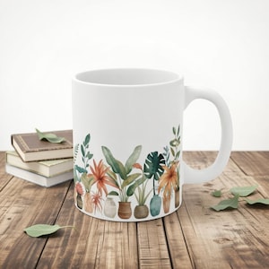 May include: White ceramic mug with a watercolour design of potted plants in green, orange, and brown. The mug has a curved handle and is on a wooden surface with books and leaves.