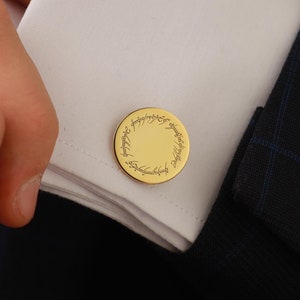 May include: Gold-colored cufflinks with a circular design and engraved text. The cufflinks are on a white dress shirt cuff, paired with a dark blue suit jacket. The image shows a close-up of the cuff and the person's hand.
