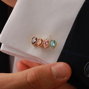 May include: A white dress shirt with gold cufflinks featuring four small, colorful gemstones. The cufflinks are in a row, with a purple, pink, light pink, and blue gemstone.