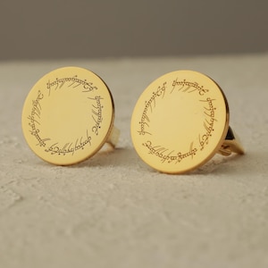 Puede incluir: Dos anillos dorados con un diseño circular y texto grabado en el borde. Los anillos están sobre un fondo neutro. El diseño y el texto recuerdan un tema de fantasía.